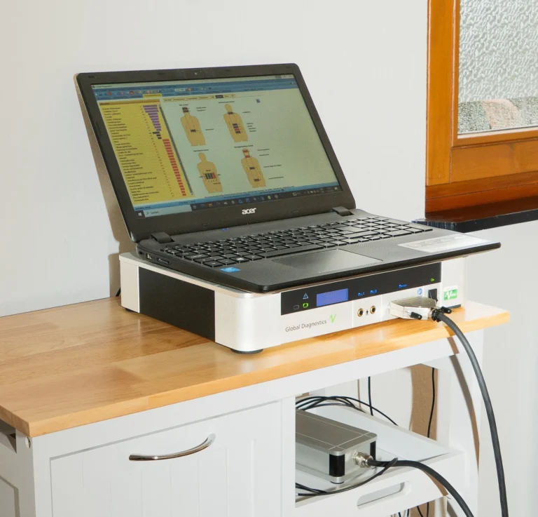 Ein schwarzer Acer-Laptop steht auf einem flachen, weiß-silbernen „Global Diagnostics“-Diagnosegerät mit Kabelanschlüssen. Auf dem Bildschirm ist eine medizinische Diagnostik-Software mit Grafiken des menschlichen Körpers und Diagrammen geöffnet. Der Aufbau steht auf einem hellen Holzschrank neben einem Fenster.