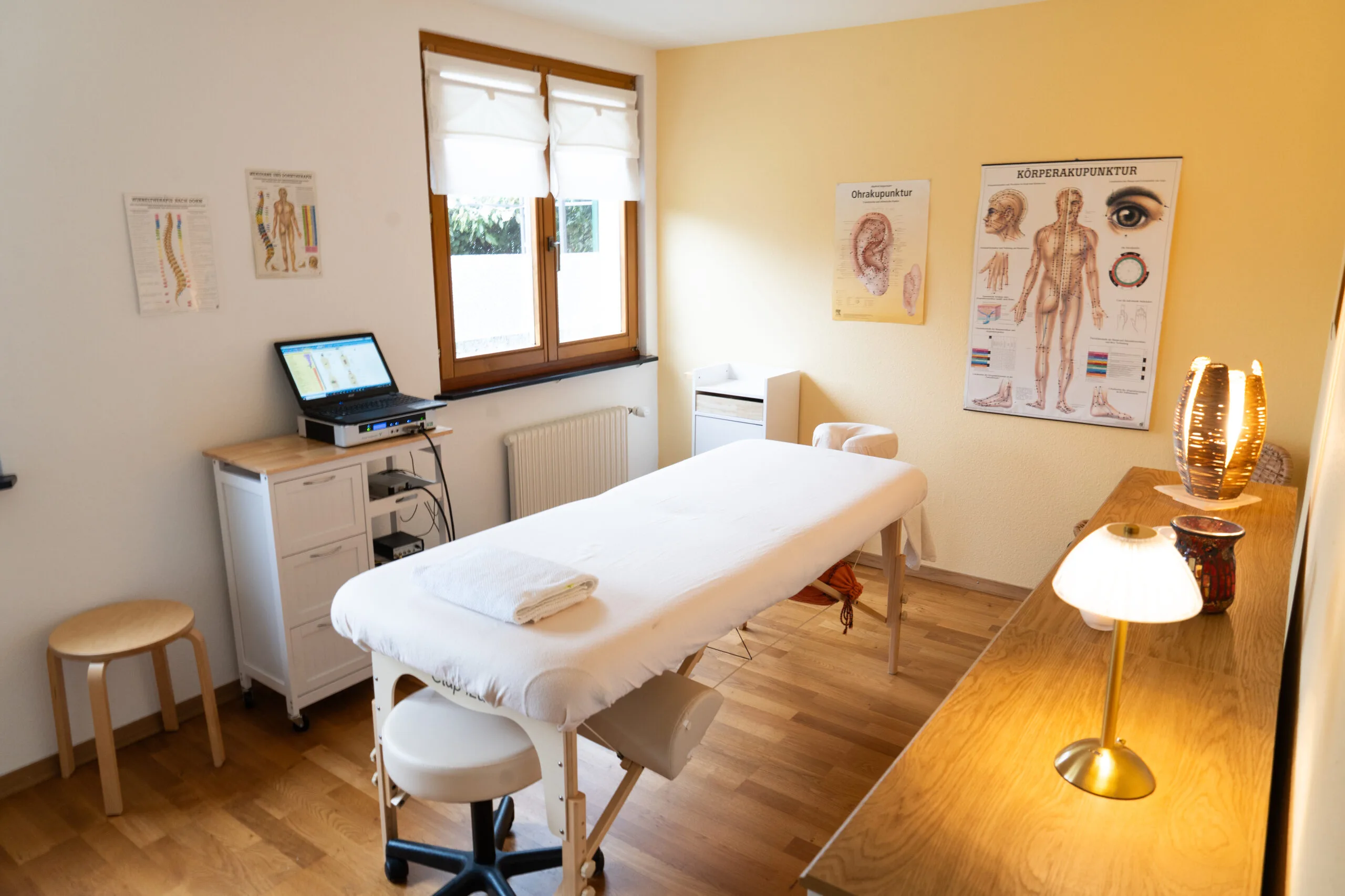 Weitwinkelaufnahme des hellen Praxisraums mit warmem Licht, Holzboden und gelber Akzentwand. In der Raummitte steht eine weiß bezogene Behandlungs- und Massageliege mit einem Rollhocker darunter. Links stehen ein Holzschemel sowie ein weißer Rollschrank mit einem Laptop zur Diagnose. An den Wänden hängen Lehrtafeln zu Ohrakupunktur, Körperakupunktur und Wirbeltherapie.