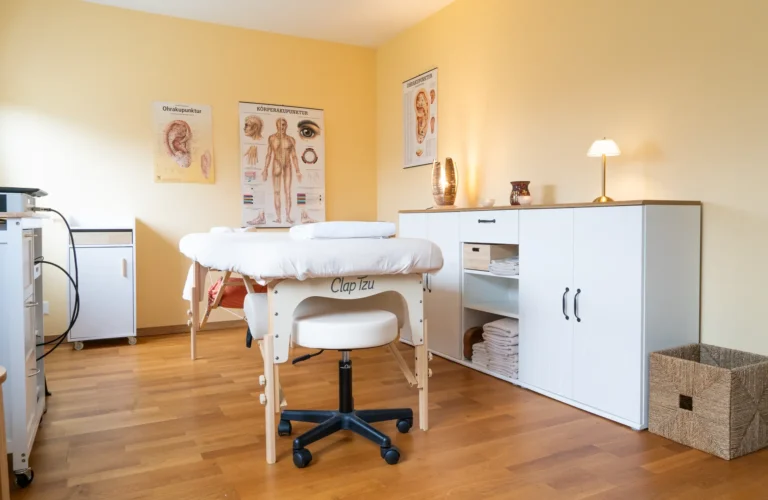 Warmer, hell gelb gestrichener Praxisraum mit Holzboden. Im Vordergrund steht eine weiß bezogene Behandlungs- und Massageliege mit einem passenden weißen Rollhocker davor. Rechts befindet sich ein weißes Sideboard mit Deko und kleinen Lampen, während an den Wänden verschiedene anatomische Lehrtafeln hängen.