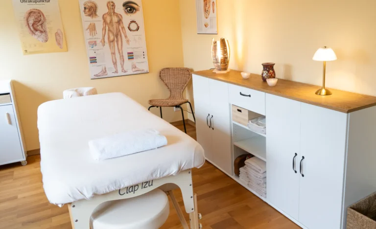 Praxisraum mit warmem Licht und gelber Wandgestaltung. Im Zentrum steht eine weiß bezogene Behandlungs- und Massageliege mit zusammengelegtem Handtuch darauf. Rechts hängen anatomische Lehrtafeln an der Wand, links steht ein Holzfenster und daneben ein Laptoptisch.