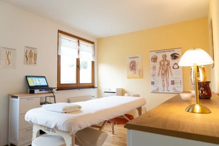 Behandlungsraum der Naturheilpraxis mit Therapieliege, Holzfenster und Anatomiepostern an der gelben Wand.